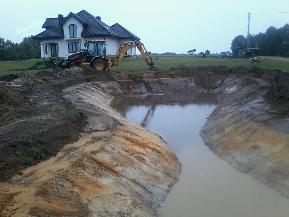 Żółta koparko-ładowarka wykonuje prace ziemne przy tworzeniu zbiornika wodnego na tle białego domu jednorodzinnego.