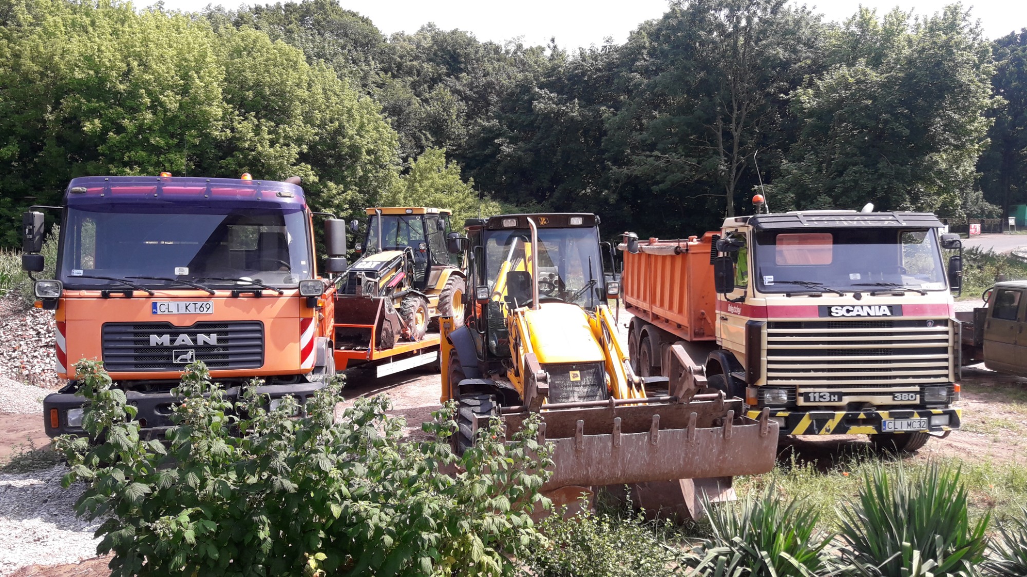 Pomarańczowy samochód ciężarowy marki MAN z fioletową szybą czołową, żółta koparko-ładowarka JCB z opuszczoną łyżką, beżowo-pomarańczowa ciężarówka Scania 113H oraz inne maszyny budowlane...