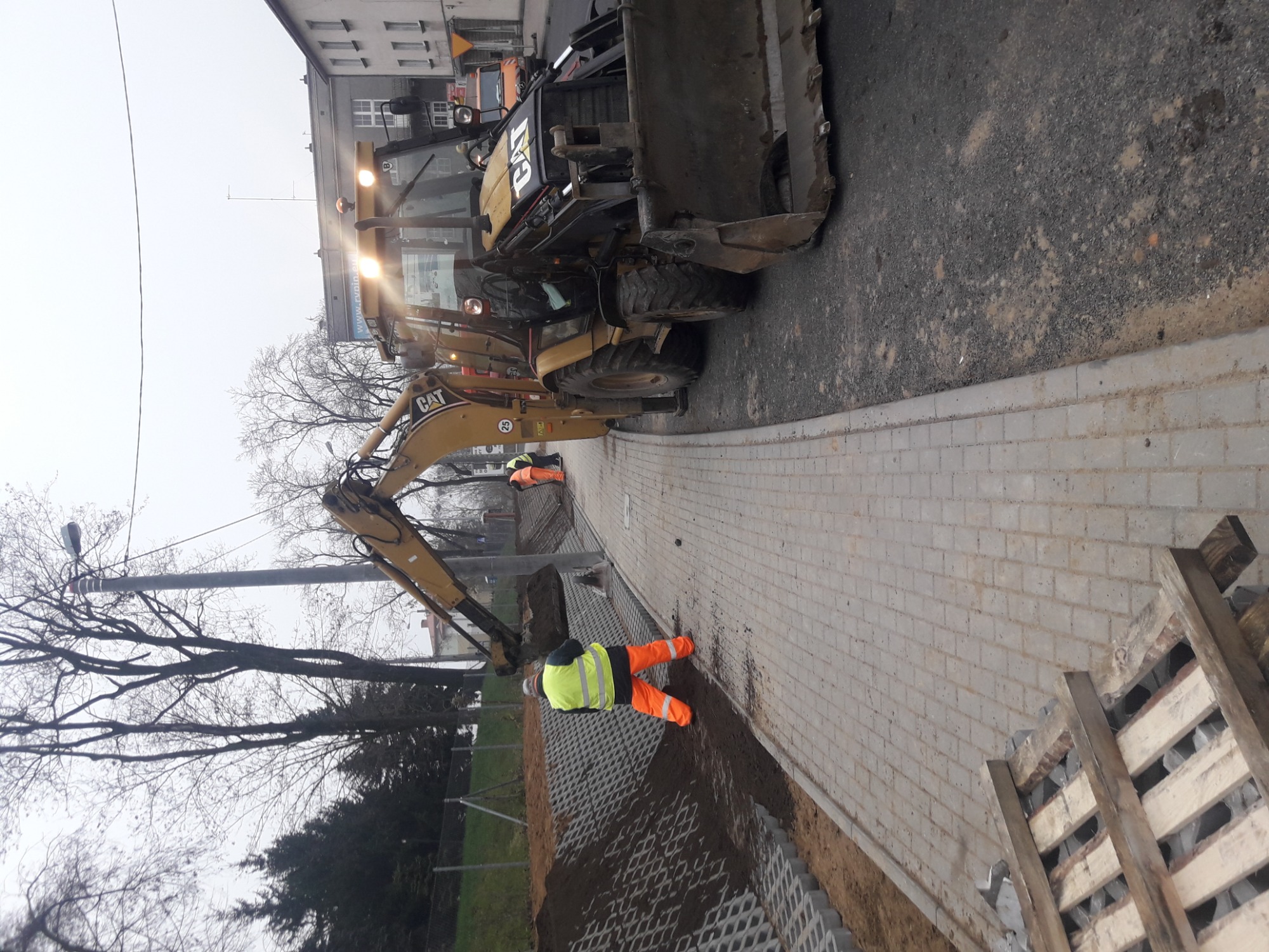 Żółta koparko-ładowarka CAT na placu budowy, pracownik w odblaskowej kamizelce przy układaniu kostki brukowej, widoczne elementy stabilizujące grunt. Ujęcie z góry, pochmurne niebo w tle.