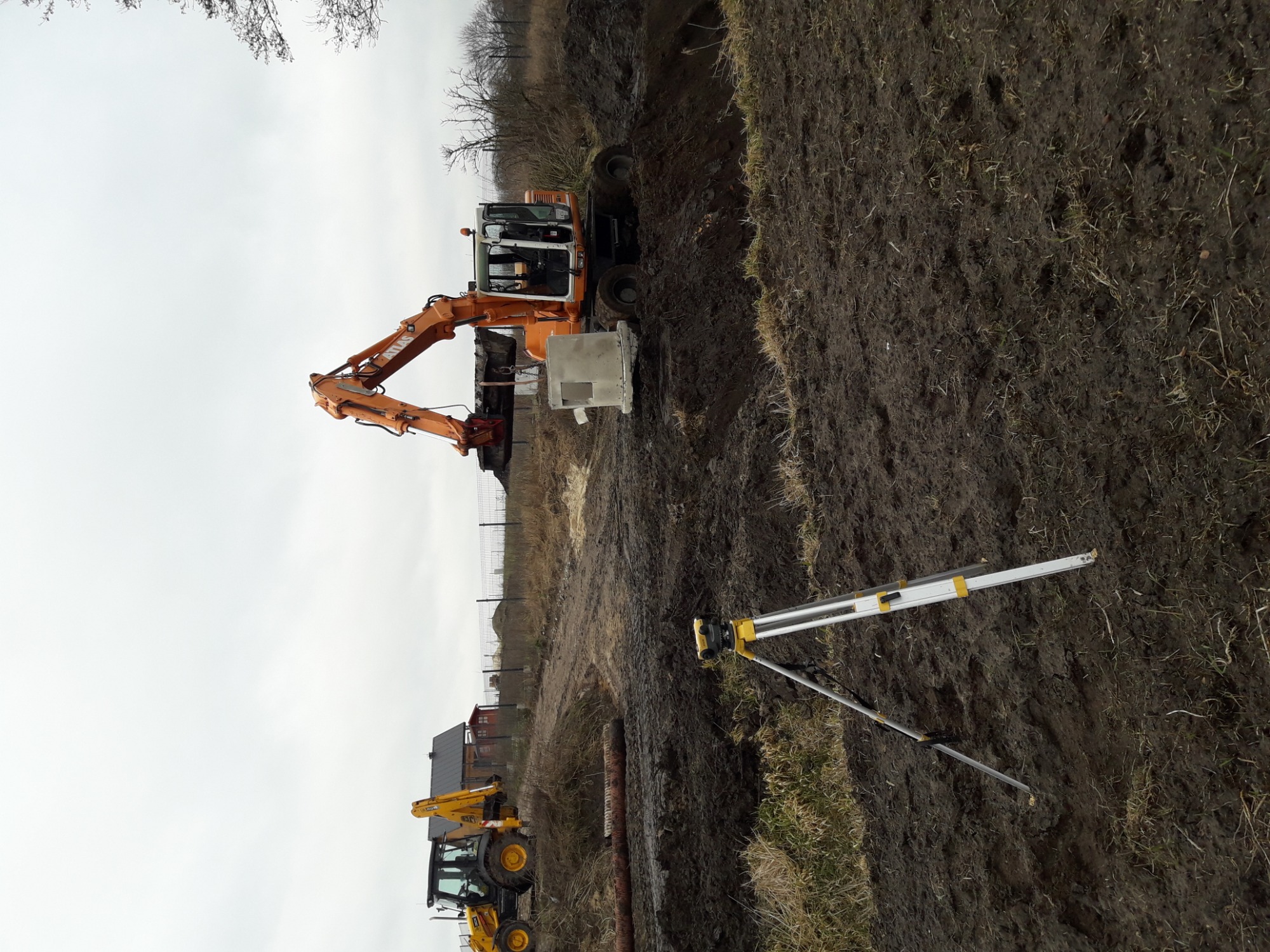 Pomarańczowa koparka z betonowym elementem w wykopie, obok niwelator na statywie i druga koparka w tle, teren pokryty błotem.