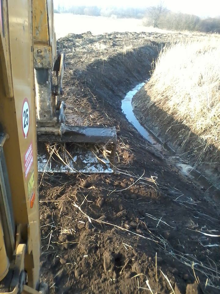 Żółta koparka w trakcie wykonywania rowu melioracyjnego, w wykopie zbiera się woda, widoczne ślady opon i świeżo wykopana ziemia.