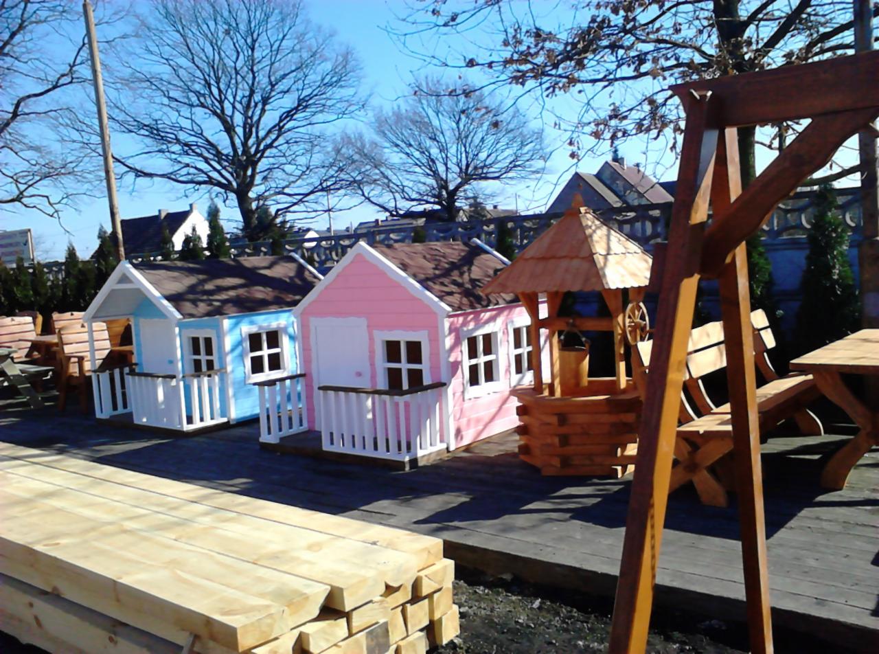Dwa kolorowe domki dla dzieci (niebieski i różowy) z białymi balustradami, drewniana studnia z wiadrem i drewniany stół z ławkami na drewnianym tarasie.
