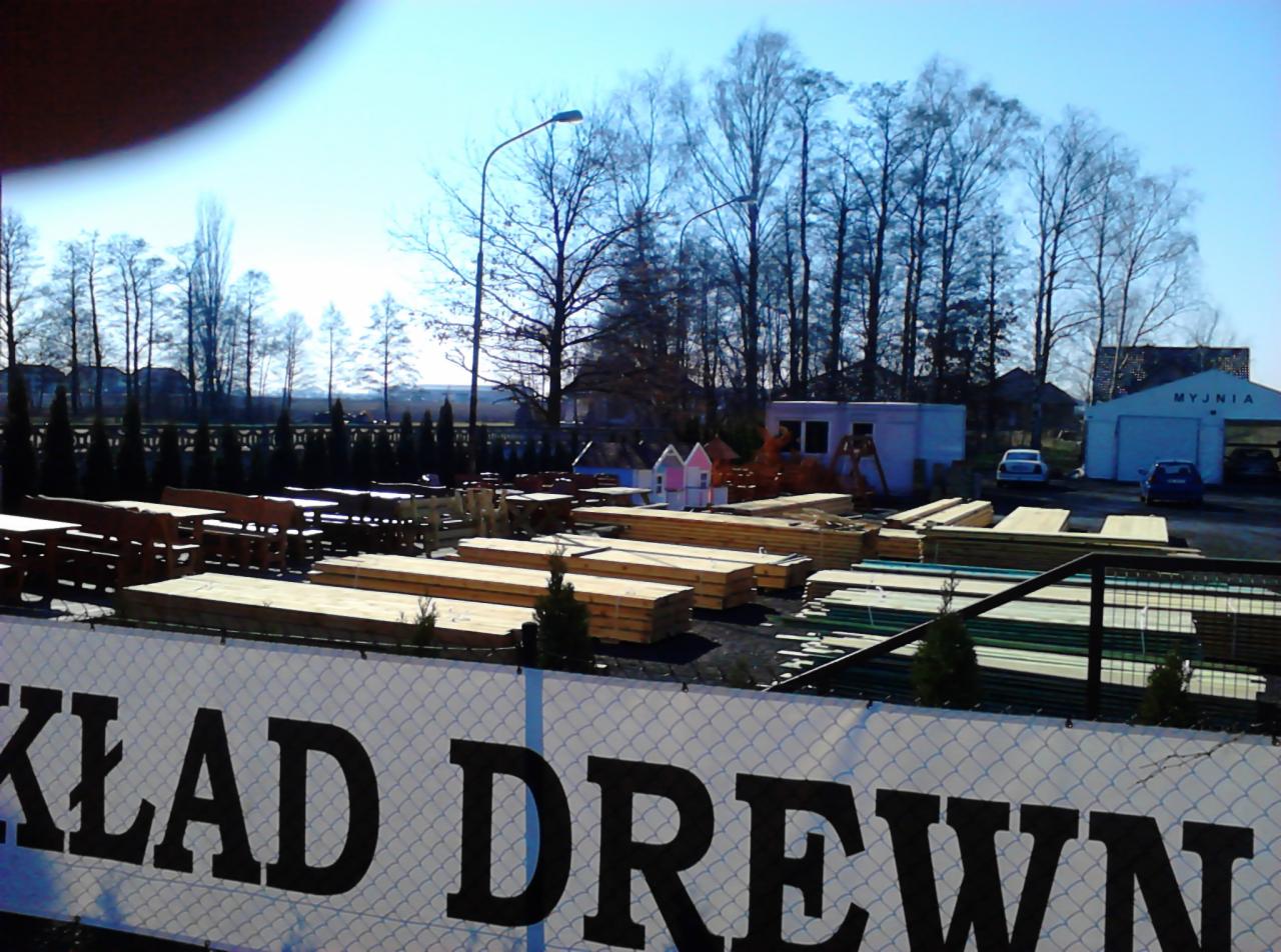 Stosy tarcicy i desek na placu, widoczne także drewniane meble ogrodowe, w tle budynki i drzewa, na pierwszym planie baner z napisem.