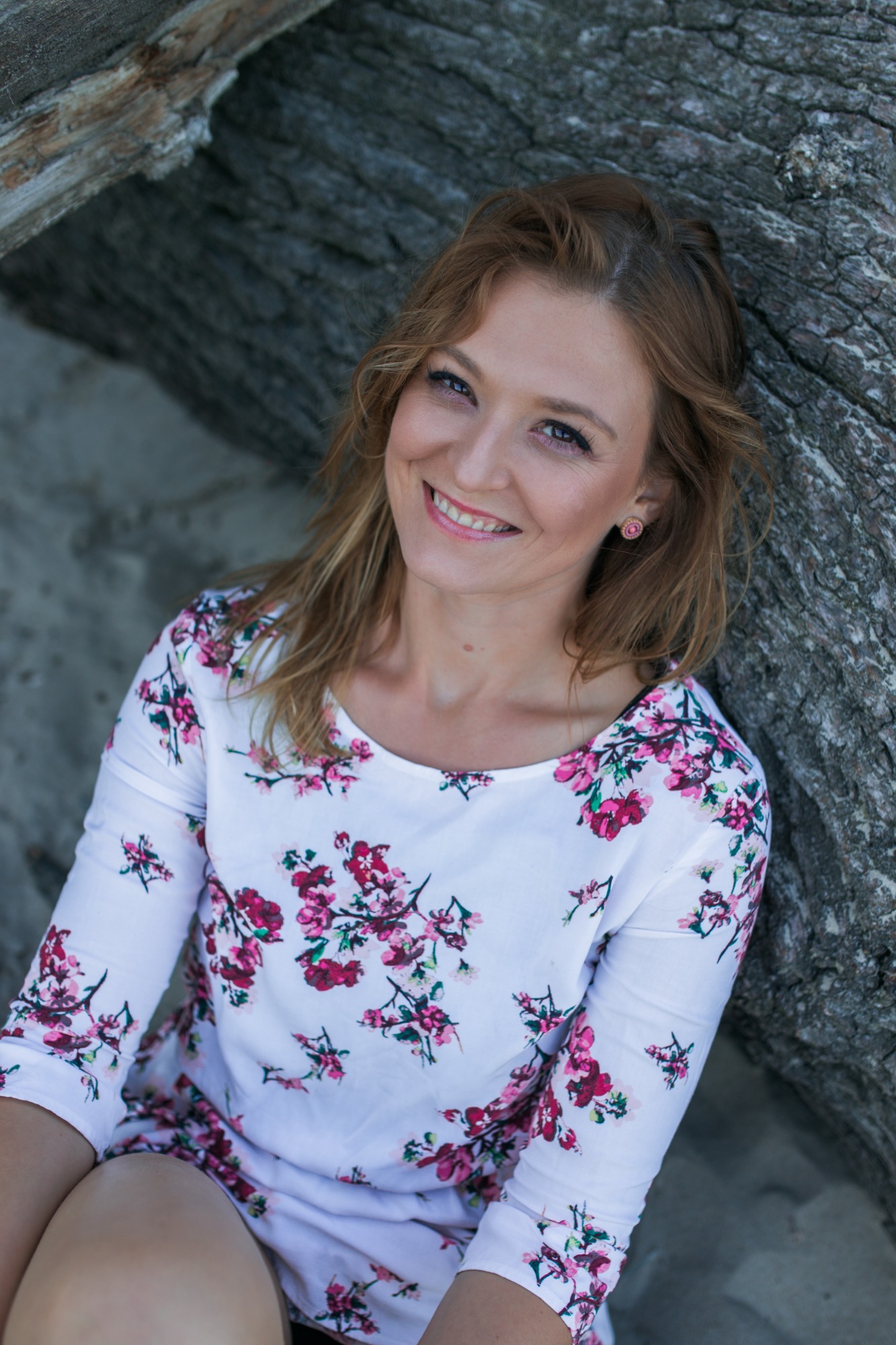 Uśmiechnięta kobieta z blond włosami, w kwiecistej sukience, oparta o szorstkie, jasne drewno, prezentuje naturalny makijaż i delikatne kolczyki.