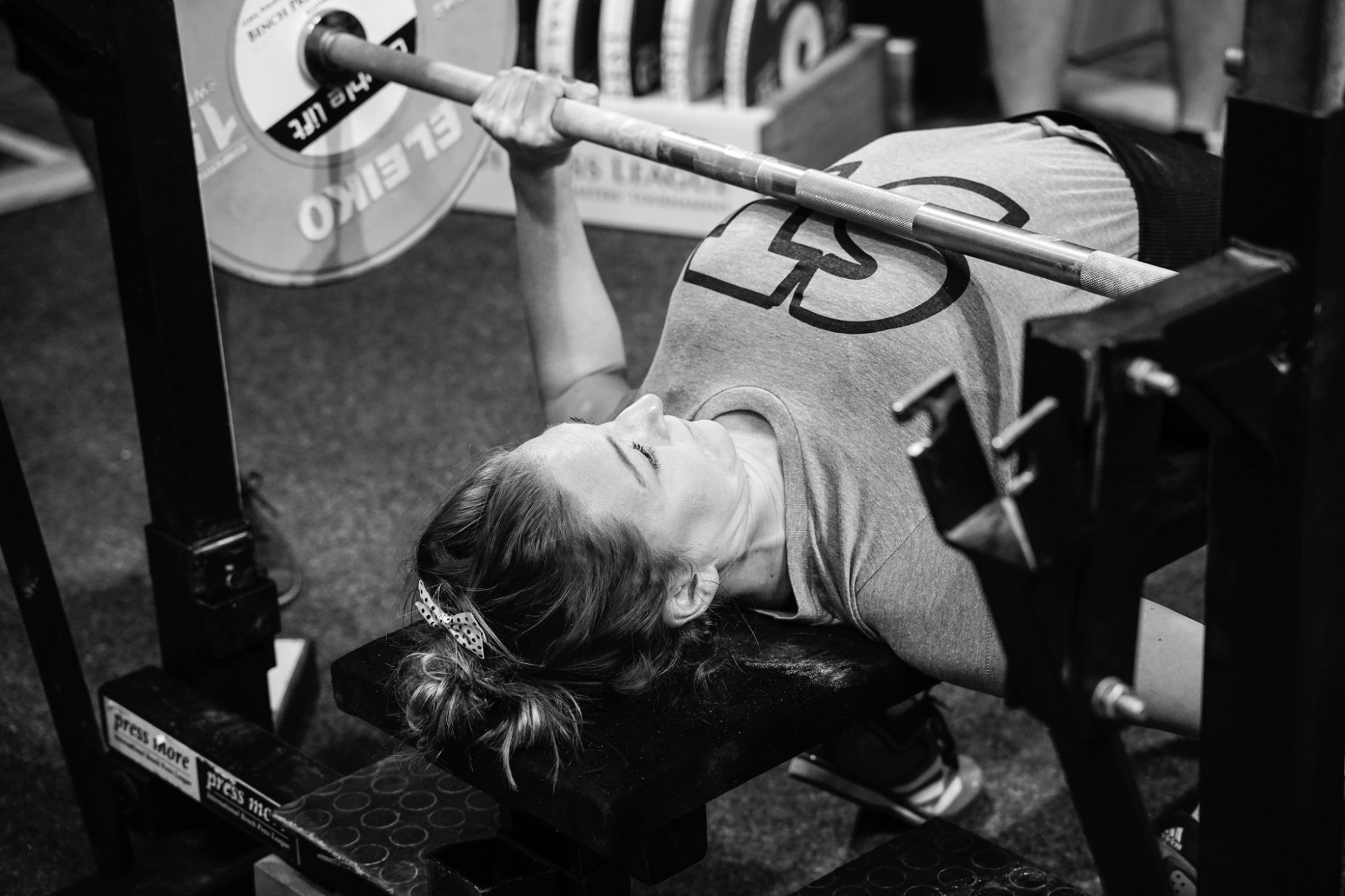 Czarno-białe zdjęcie kobiety leżącej na ławce i podnoszącej sztangę na siłowni, widoczny ciężar marki Eleiko, kobieta ma włosy upięte spinką w kokardę.
