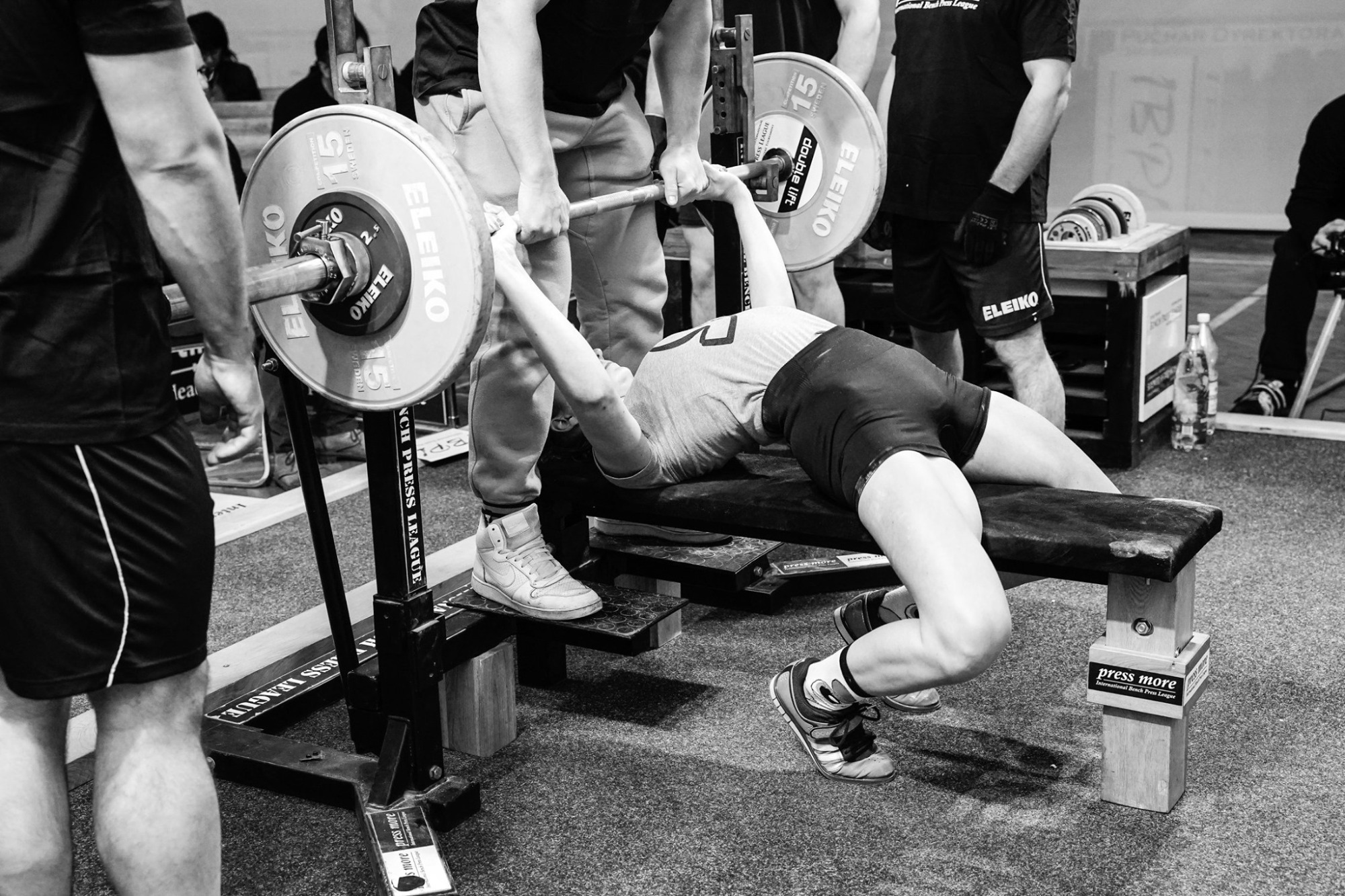 Czarno-białe zdjęcie osoby ćwiczącej wyciskanie sztangi leżąc, z asekuracją dwóch osób, na tle siłowni z widocznymi ciężarami i sprzętem sportowym.