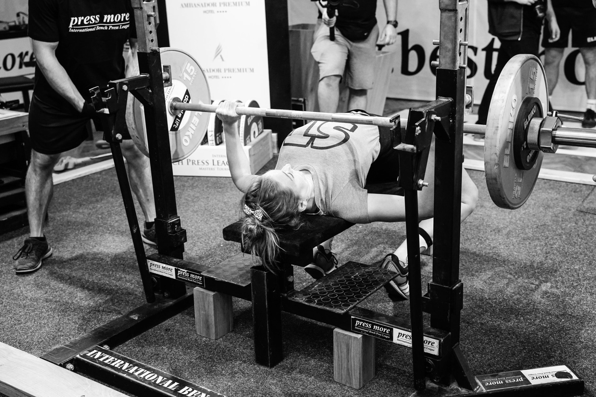 Kobieta w koszulce z numerem 45 wyciska sztangę na ławce podczas zawodów International Bench Press League, asystowana przez mężczyznę w czarnej koszulce, w tle widoczny baner reklamowy hotelu...