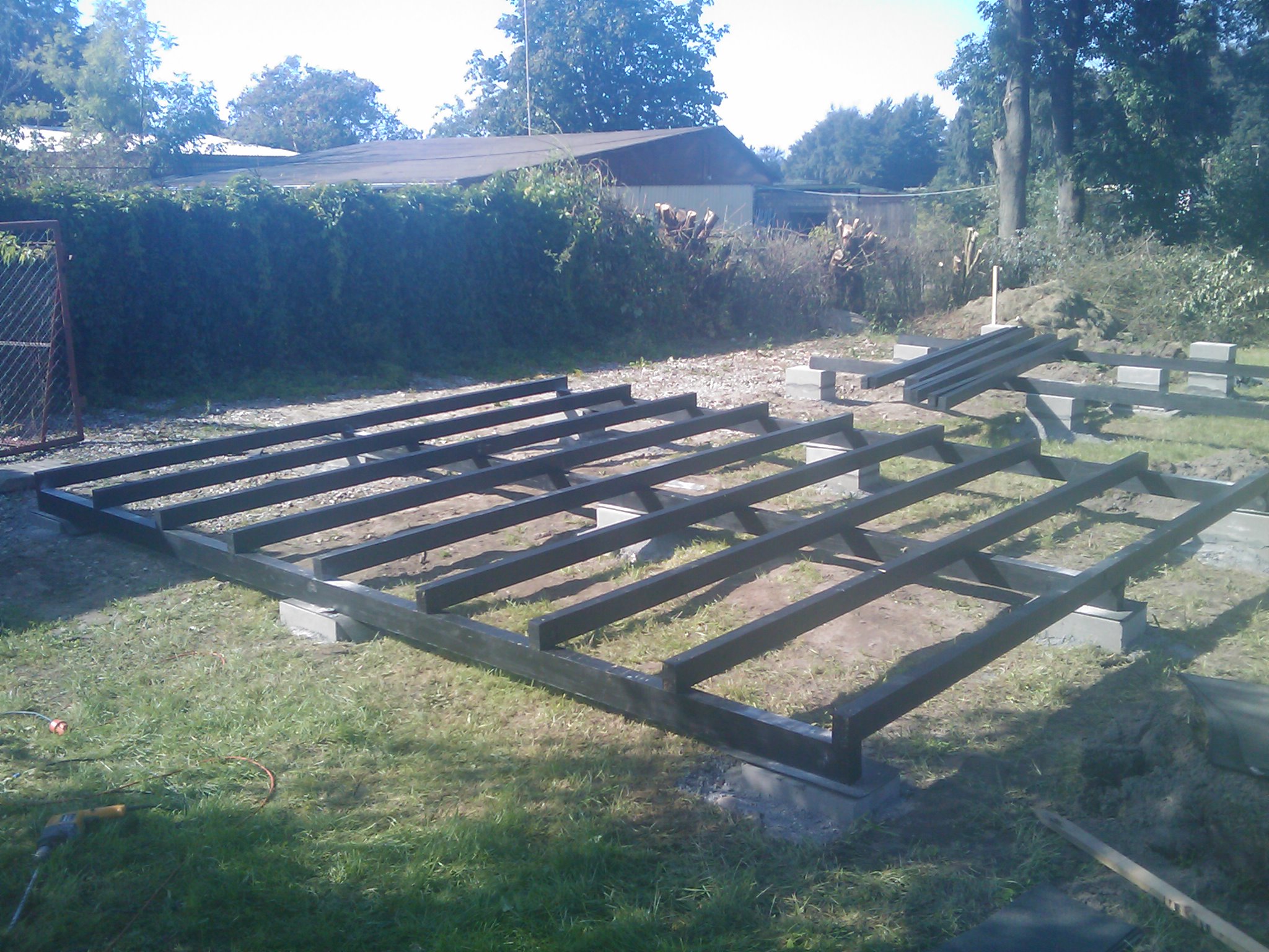 Drewniana konstrukcja pod dom letniskowy, osadzona na betonowych bloczkach, widoczna na trawniku z zielonym żywopłotem w tle.