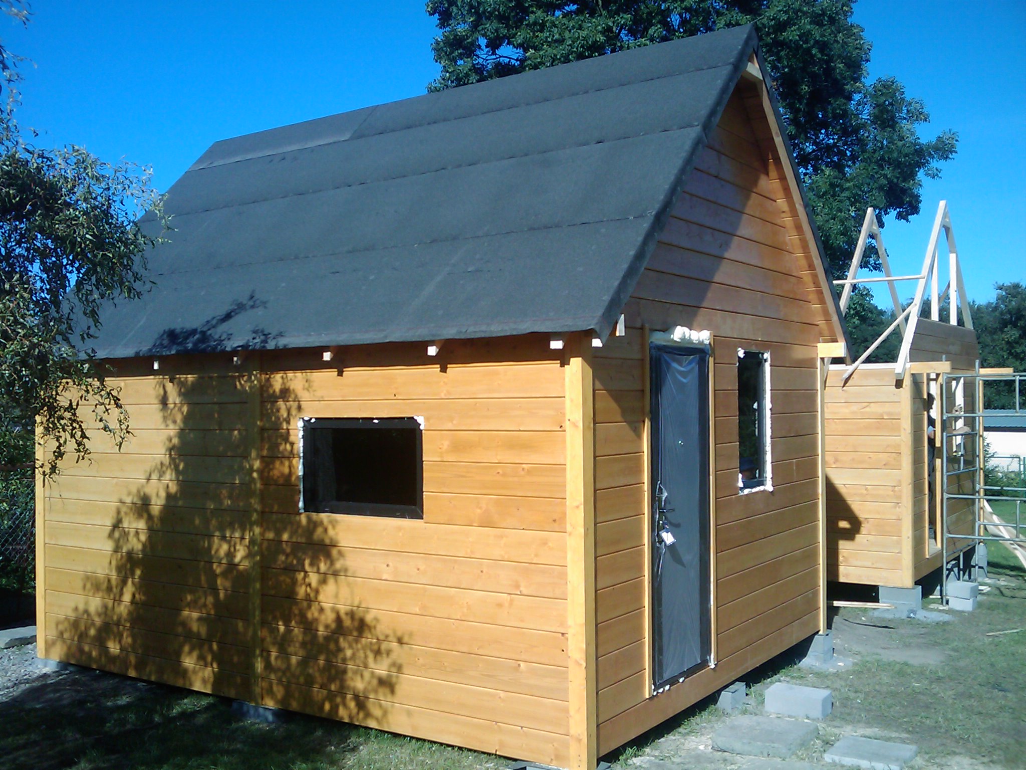Drewniany domek letniskowy w Jankowie Przygodzkim z czarnym dachem z papy, widoczne okno i drzwi zabezpieczone folią, obok drugi domek w trakcie budowy.