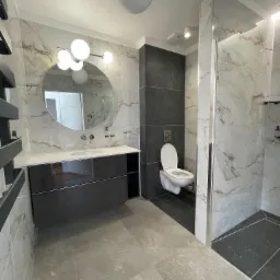 Nowoczesna łazienka z marmurowymi płytkami, okrągłym lustrem z oświetleniem i podwieszaną toaletą na tle ciemnoszarej ściany.