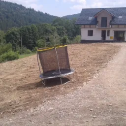 Teren po robotach ziemnych z niedokończonym domem w tle i trampoliną. Widoczna droga dojazdowa.