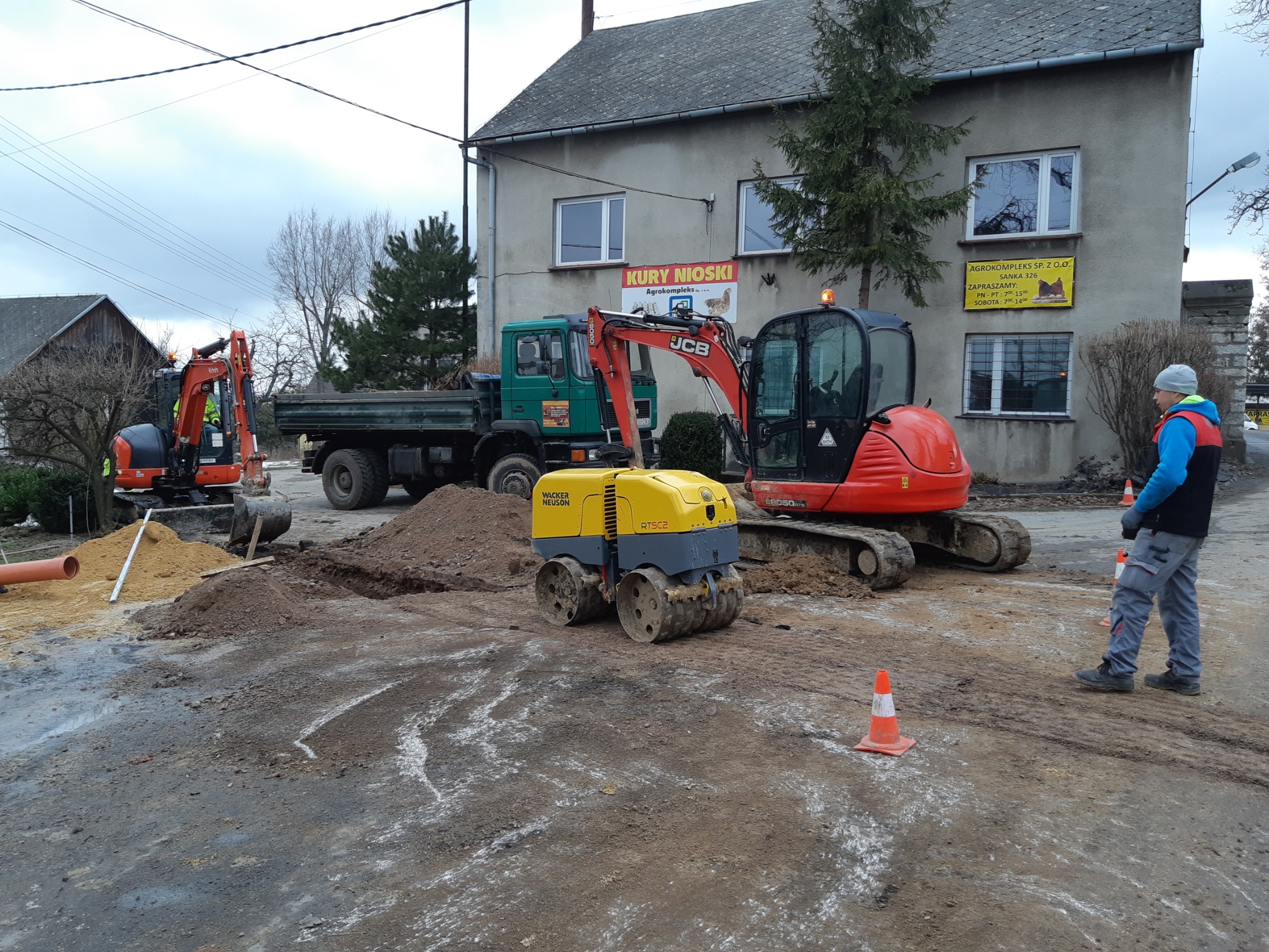 Przygotowanie podłoża pod drogę: walec Wacker Neuson, koparka JCB i Kubota oraz wywrotka na tle budynku z reklamą Kury Nioski.