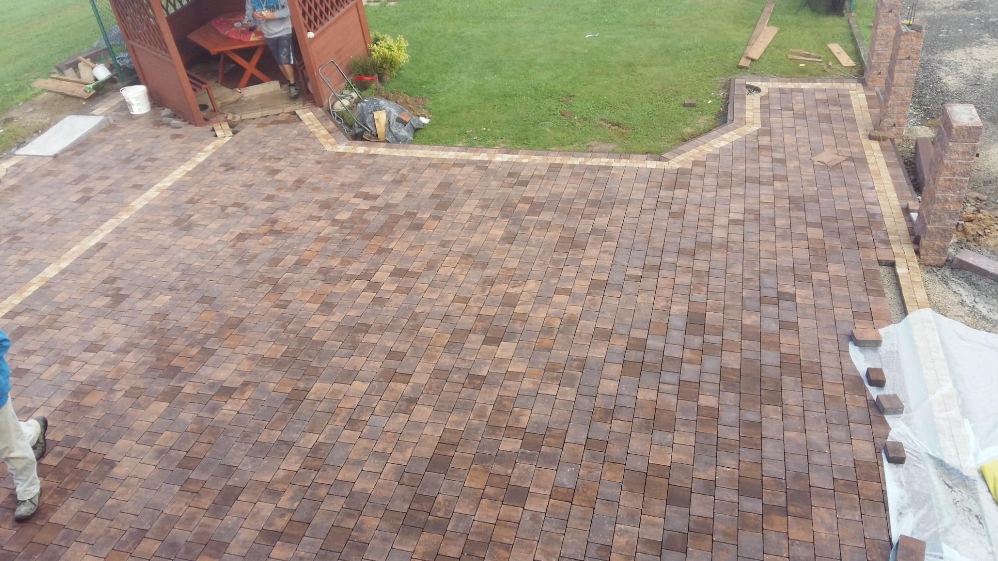 Układanie kostki brukowej w odcieniach brązu i beżu, widok z góry na ułożoną nawierzchnię z obrzeżami, w tle altana, trawnik i częściowo ułożone słupki ogrodzeniowe.