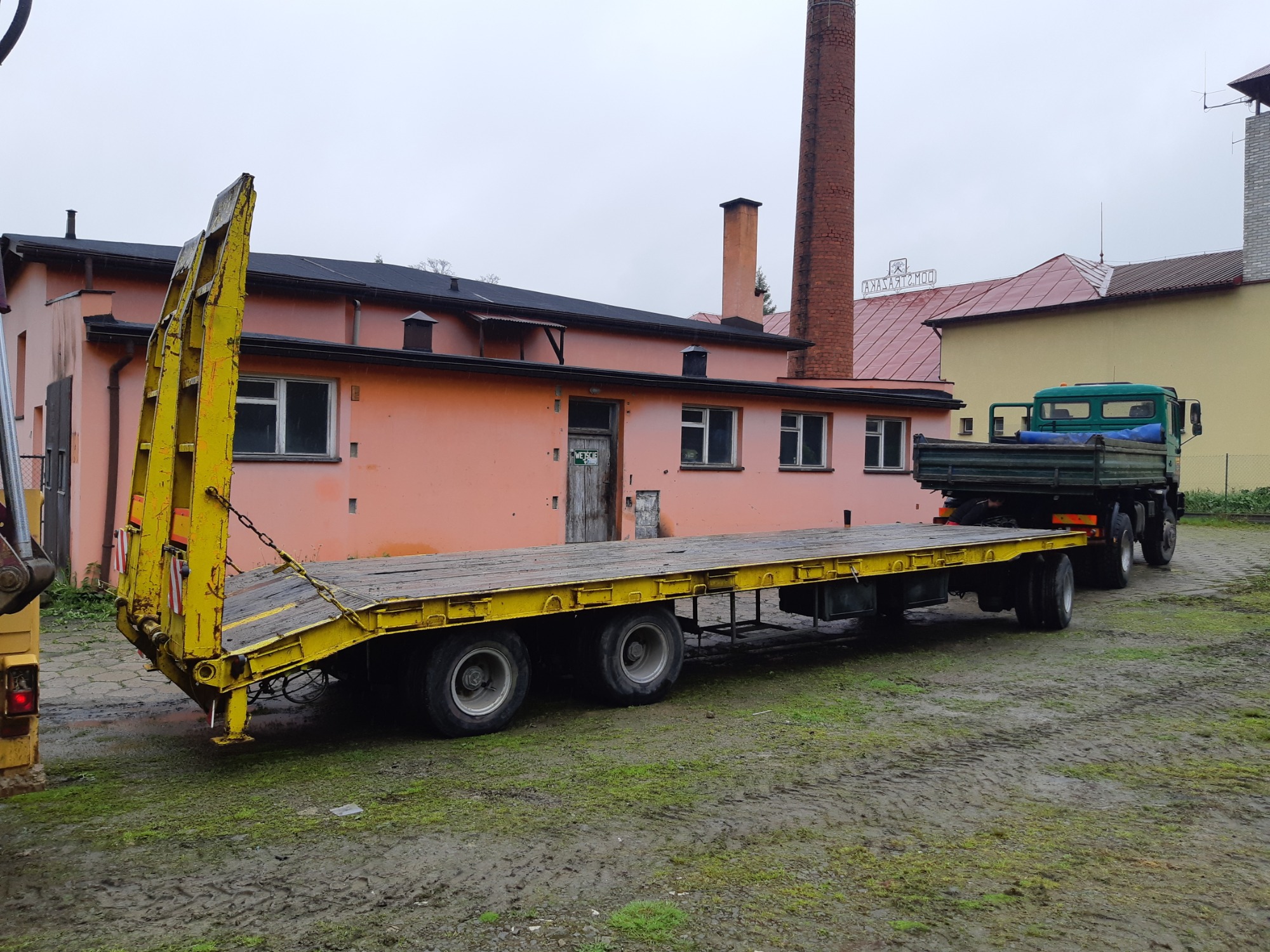 Żółta naczepa niskopodwoziowa na błotnistym terenie, z zieloną ciężarówką w tle i budynkami z ceglanym kominem.