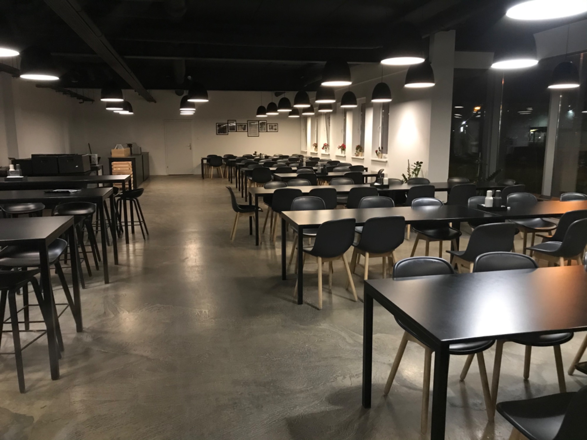 Sala konferencyjna lub stołówka biurowa z rzędami czarnych stołów i krzeseł, oświetlona wiszącymi lampami. Na parapecie okna doniczki z kwiatami.