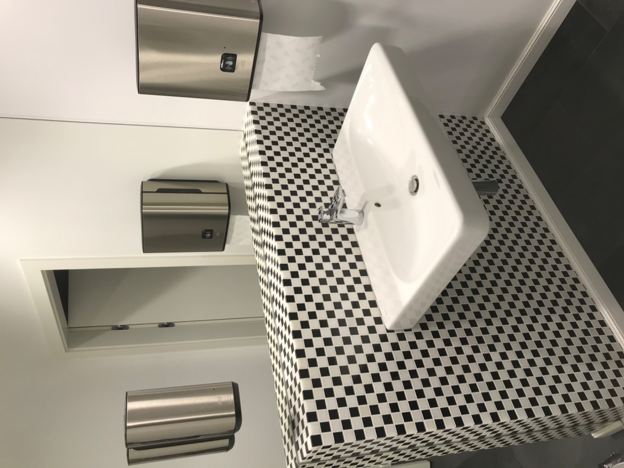 Biała umywalka z chromowaną baterią na blacie wyłożonym czarno-białą mozaiką w publicznej toalecie, obok metalowy podajnik na ręczniki papierowe i dozownik mydła.