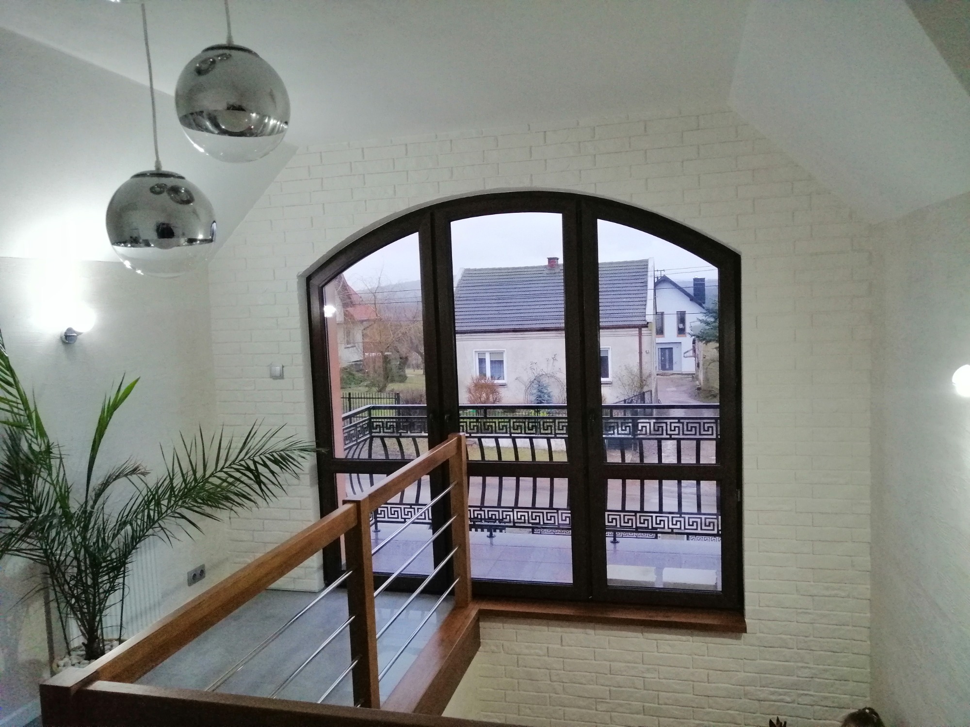 Wnętrze domu w Mniowie z białymi ścianami imitującymi cegłę, dużym oknem łukowym z widokiem na zewnątrz i drewnianą balustradą schodów.