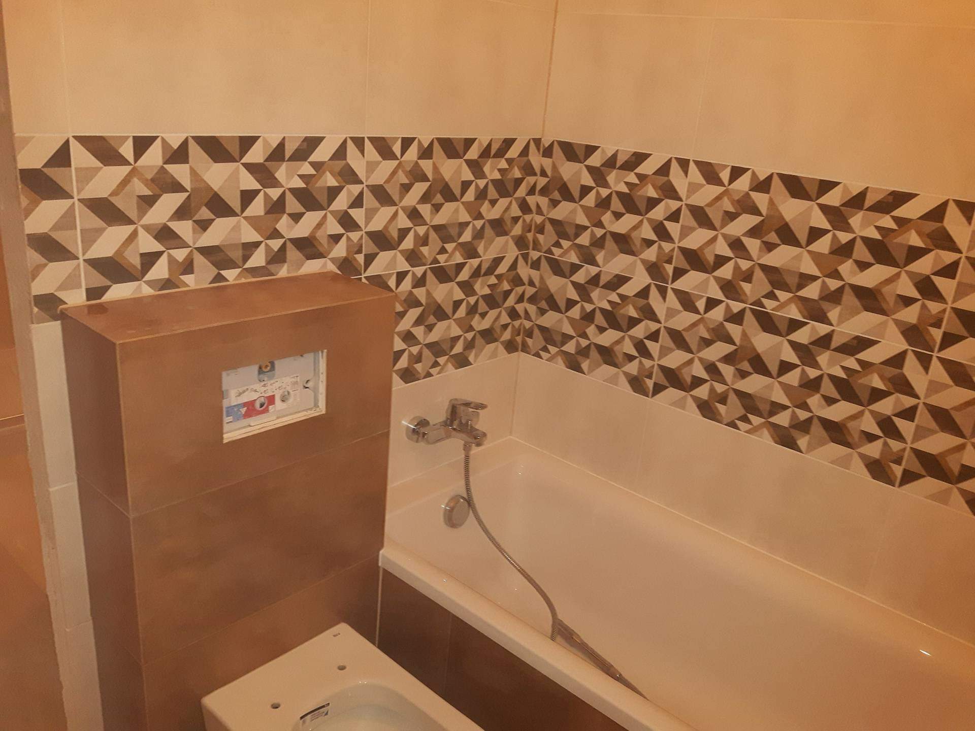 Wykończona łazienka z beżowymi i brązowymi płytkami o geometrycznym wzorze, zabudowana spłuczka WC i wanna z chromowaną baterią.