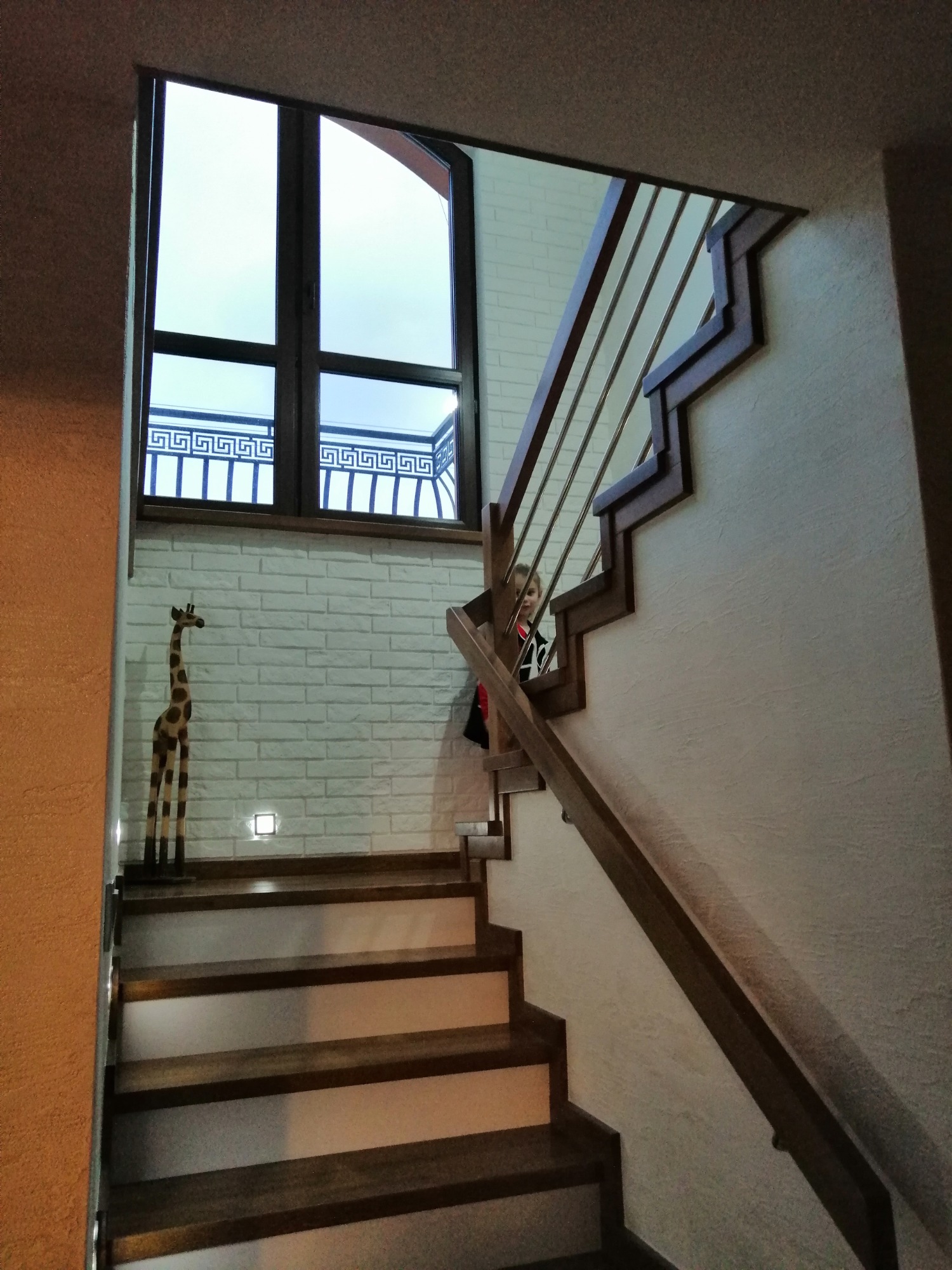 Wnętrze domu z drewnianymi schodami z podświetleniem, balustradą z metalowymi prętami i drewnianą poręczą, obok biała ściana z cegieł z drewnianą figurką żyrafy i oknem z ozdobną kratą, widoczna...