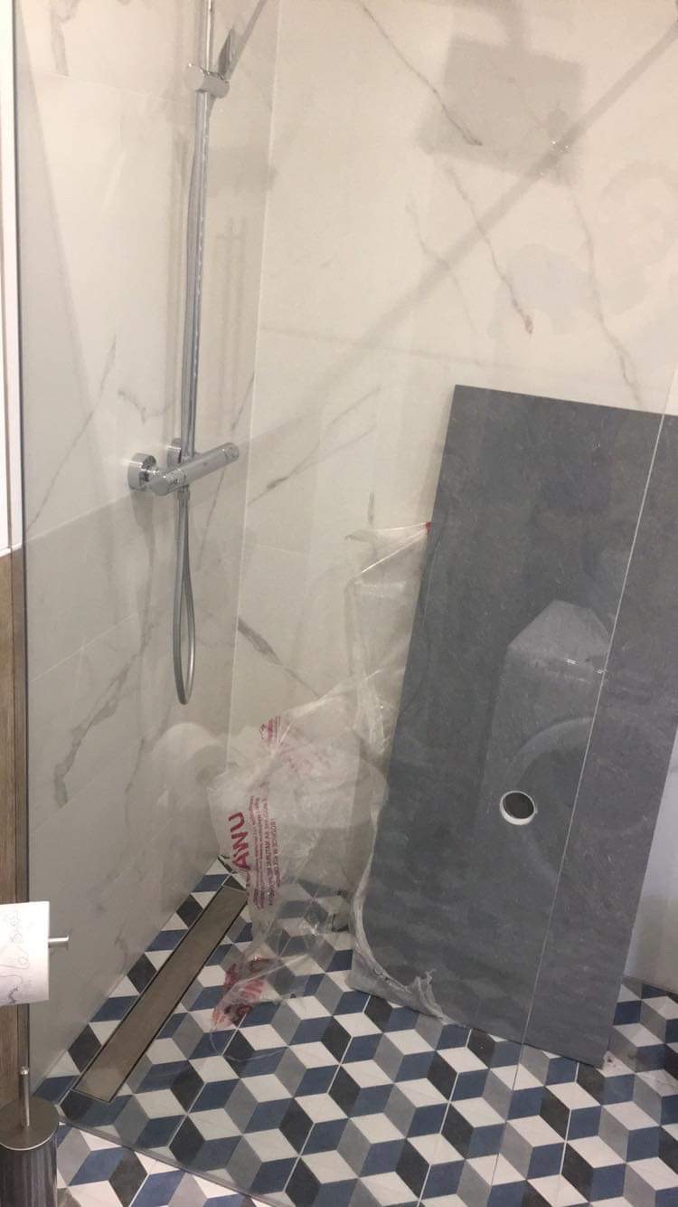 Fragment łazienki w trakcie prac wykończeniowych: zamontowany panel prysznicowy, podłoga w geometryczne wzory, płytka z otworem oparta o ścianę, folia ochronna.