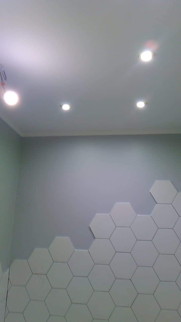 Ściana łazienki z częściowo ułożonymi białymi, heksagonalnymi płytkami, widoczne cztery punktowe lampy sufitowe i szare ściany.