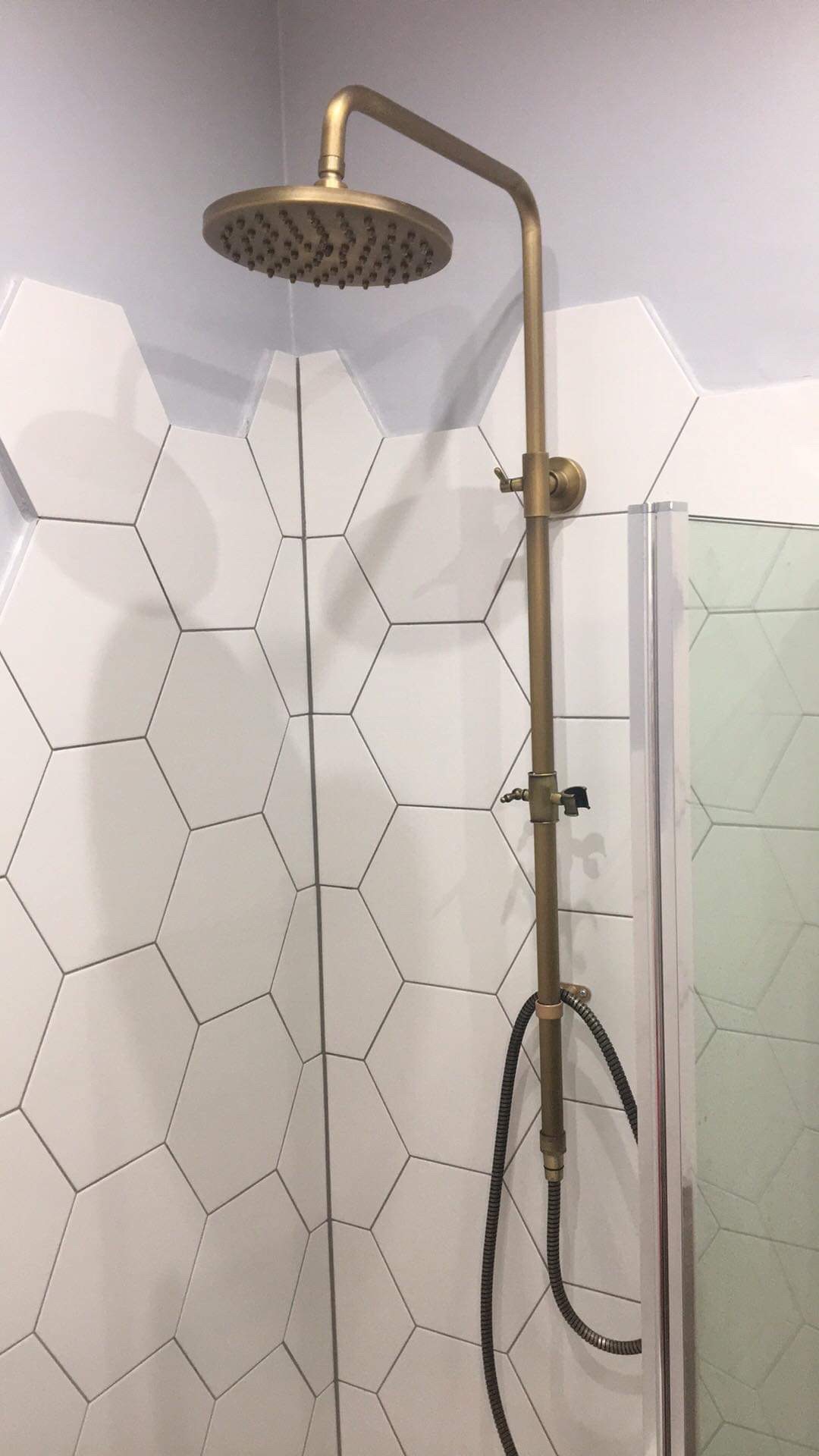 Mosiężna deszczownica z widocznymi dyszami na tle białych, heksagonalnych płytek w łazience z kabiną prysznicową.