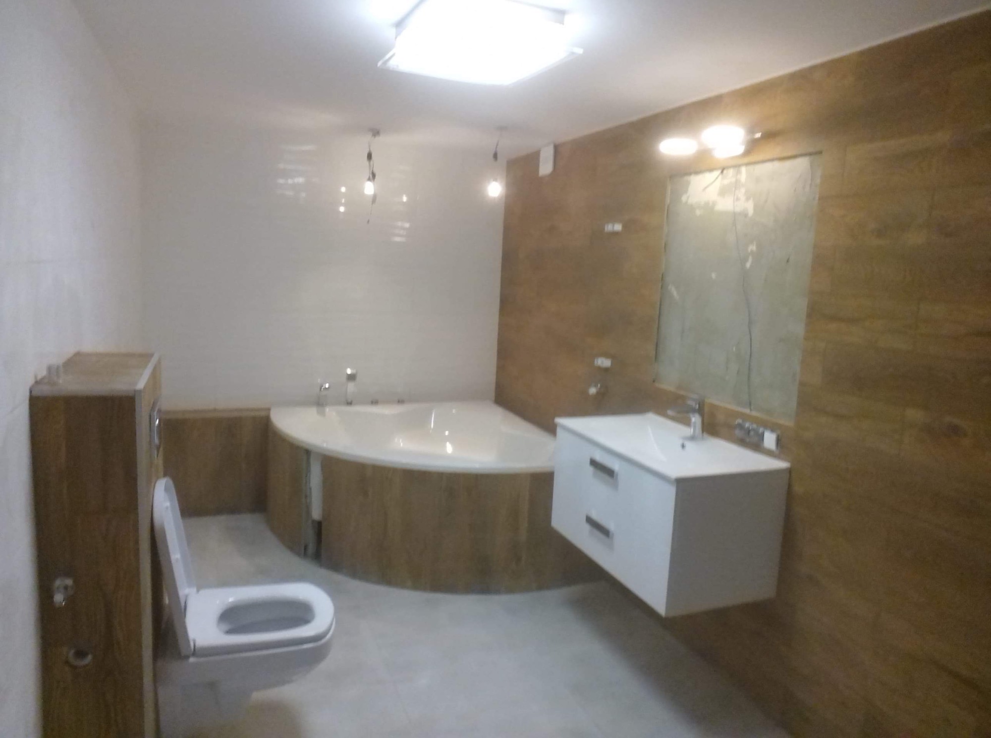 Nowoczesna łazienka z narożną wanną obudowaną drewnopodobnymi panelami, białą wiszącą szafką z umywalką i podwieszanym WC z zabudową, oświetlona lampami sufitowymi.