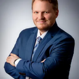 Portret uśmiechniętego mężczyzny w niebieskim garniturze i krawacie w kratę, z założonymi rękami na tle szarego gradientu.