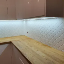 Kuchnia z drewnianym blatem i białymi płytkami w kształcie rybiej łuski, podświetlonymi listwą LED, widoczny otwór w ścianie na gniazdko.