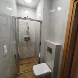 Nowoczesna łazienka z przeszkloną kabiną prysznicową, drewnopodobną podłogą, wiszącą toaletą i szarą płytką na ścianach.