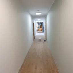 Długi korytarz po malowaniu na jasny kolor, z panelami podłogowymi imitującymi drewno, widoczny kubeł z farbą i rękawice malarskie.