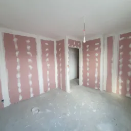 Pokój w trakcie prac wykończeniowych: ściany pokryte płytami kartonowo-gipsowymi, z widocznymi łączeniami wymagającymi szpachlowania, surowa betonowa podłoga, pojedyncza żarówka zwisająca z sufitu.