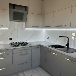 Fach-Bud - Szara, nowoczesna kuchnia z marmurowym blatem i białymi szafkami. Czarna bateria i płyta grzewcza. Oświetlenie LED pod szafkami.