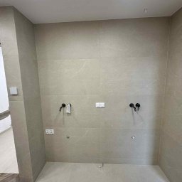 Fach-Bud - Beżowe, matowe płytki na ścianie z czarną armaturą i gniazdkami. Widoczny fragment instalacji elektrycznej i hydraulicznej. Prace wykończeniowe w toku.