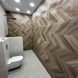 Fach-Bud - Nowoczesna łazienka z drewnopodobnymi płytkami ułożonymi w jodełkę na ścianach i szarą podłogą. Widoczna toaleta i deszczownica.