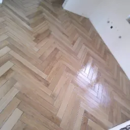 Ułożona w jodełkę, błyszcząca podłoga z płytek imitujących drewno, widok z góry na fragment pomieszczenia z białymi ścianami i widocznymi gniazdkami elektrycznymi.