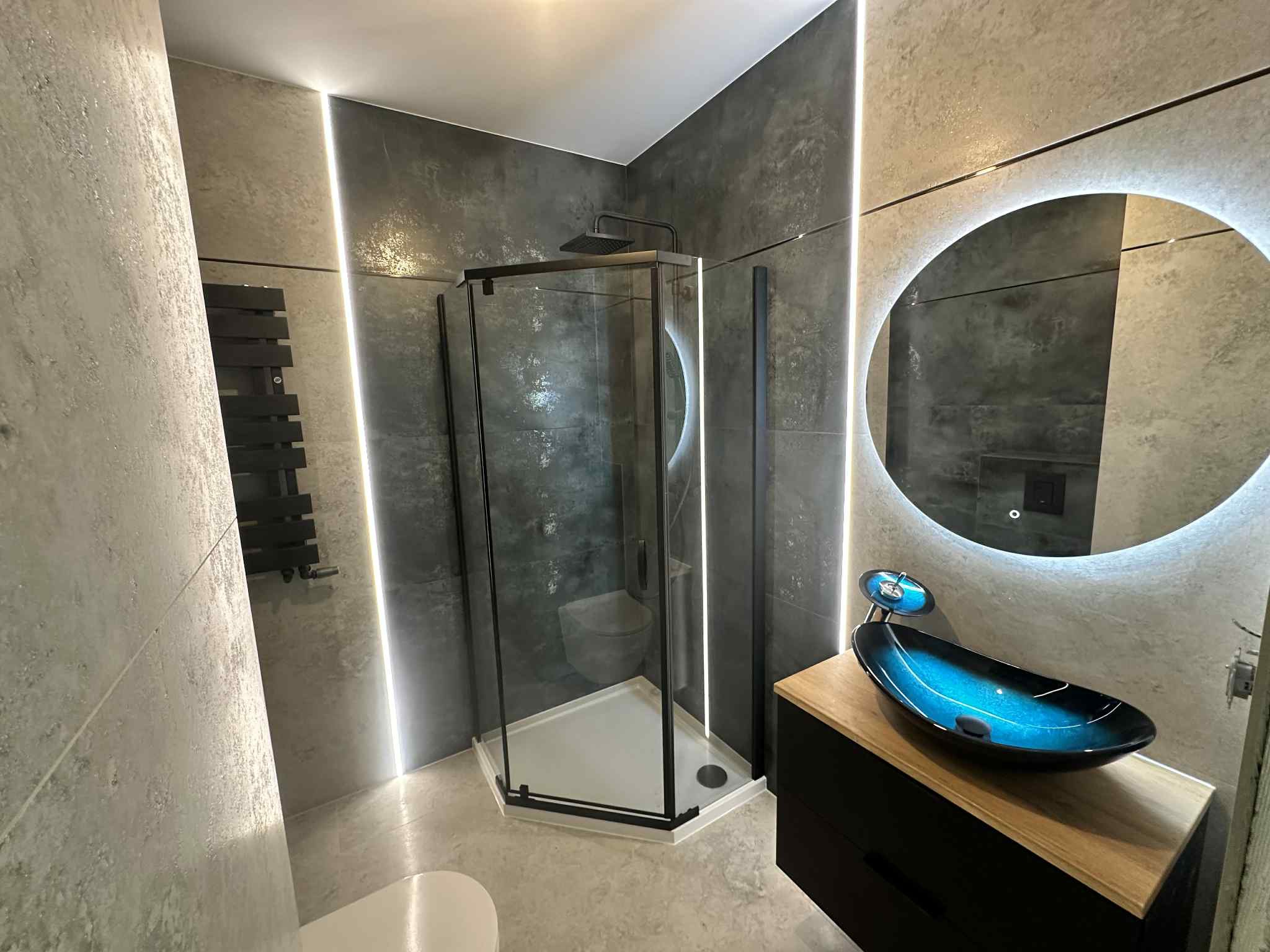 Nowoczesna łazienka z kabiną prysznicową w czarnej ramie, umywalką w odcieniach niebieskiego i okrągłym lustrem z podświetleniem LED.