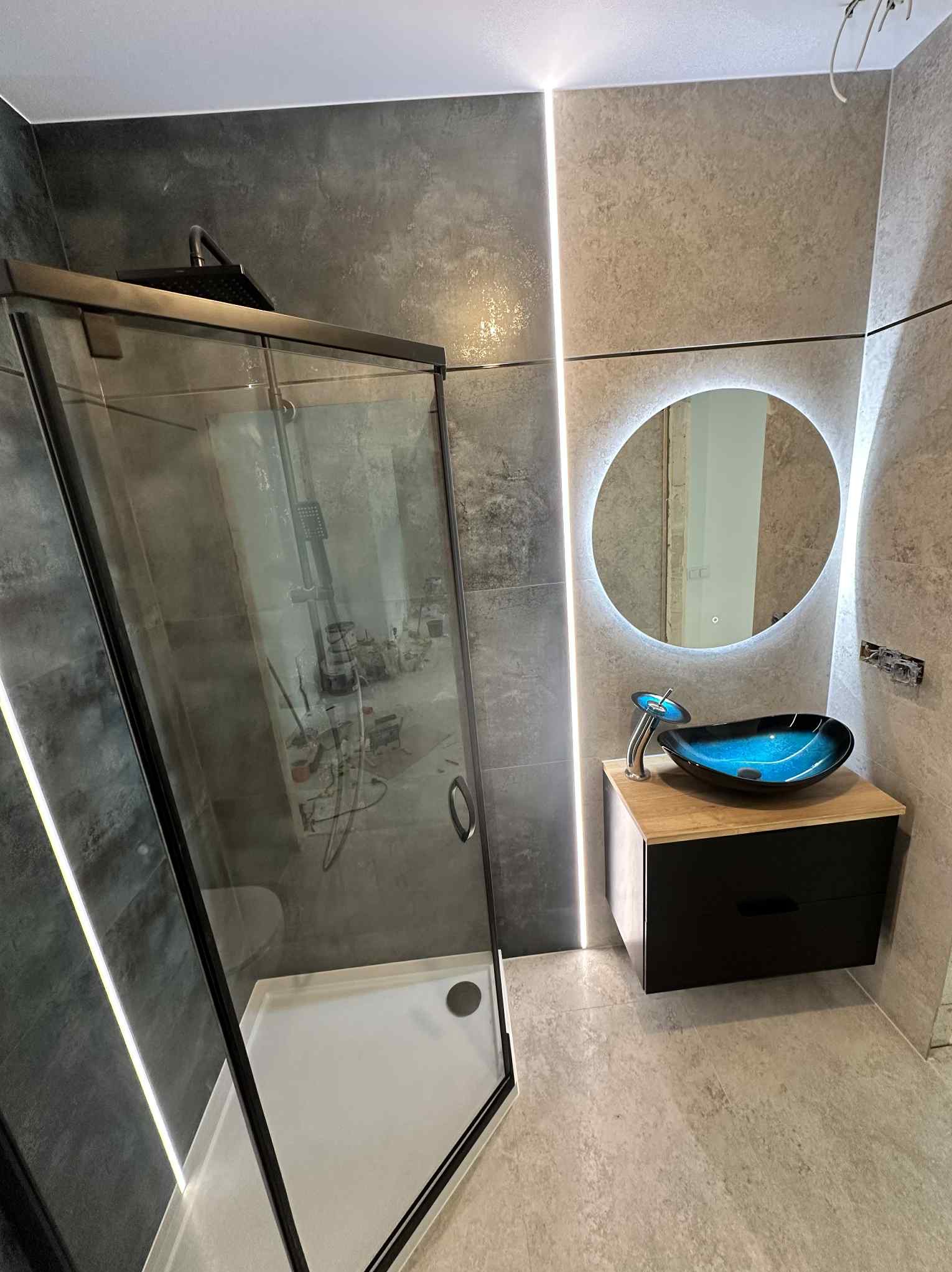 Nowoczesna łazienka z kabiną prysznicową typu walk-in, umywalką w formie misy i okrągłym lustrem z podświetleniem LED. Minimalistyczny design i eleganckie wykończenie.