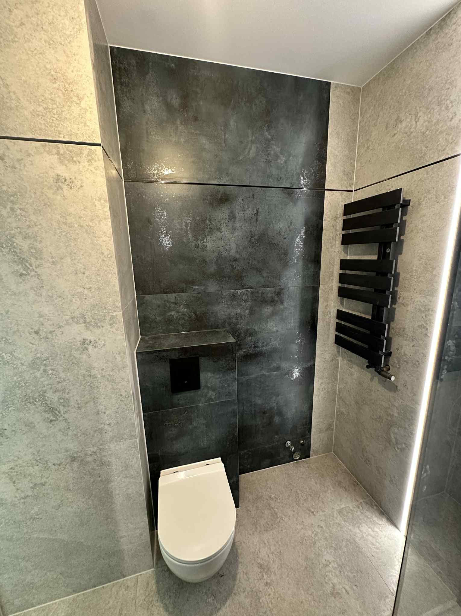 Nowoczesna łazienka z szarymi i czarnymi płytkami imitującymi beton, podwieszana toaleta i czarny grzejnik drabinkowy. Oświetlenie LED podkreśla detale.