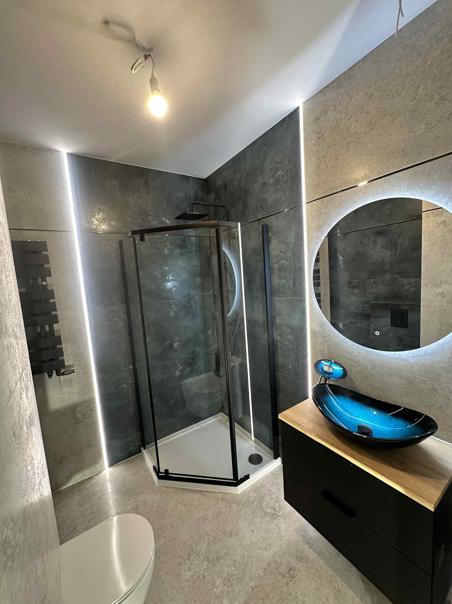 Nowoczesna łazienka z szarą armaturą, oświetleniem LED, kabiną prysznicową typu walk-in i umywalką w odcieniach niebieskiego na drewnianym blacie.