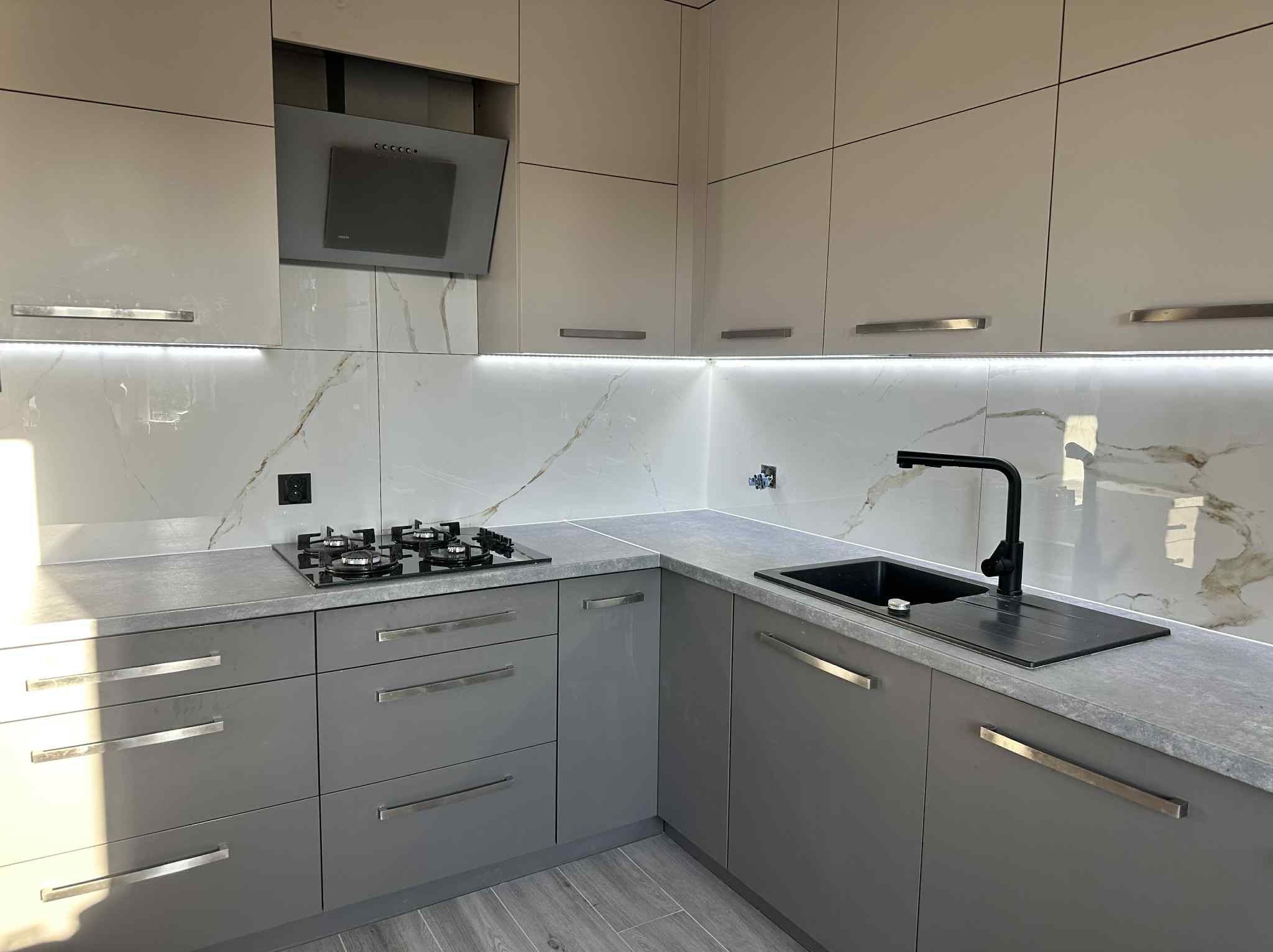 Szara, nowoczesna kuchnia z marmurowym blatem i białymi szafkami. Czarna bateria i płyta grzewcza. Oświetlenie LED pod szafkami.