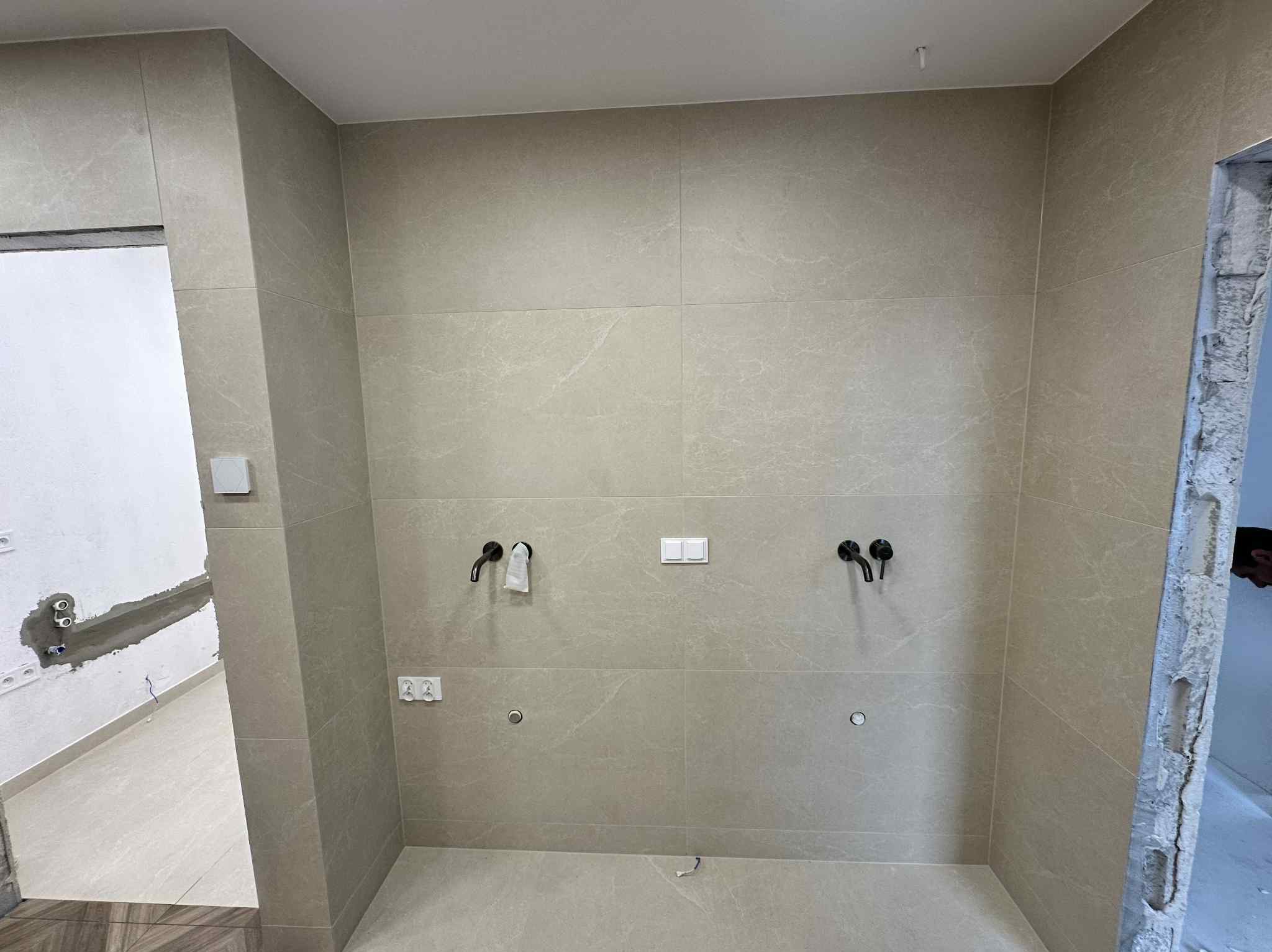 Beżowe, matowe płytki na ścianie z czarną armaturą i gniazdkami. Widoczny fragment instalacji elektrycznej i hydraulicznej. Prace wykończeniowe w toku.