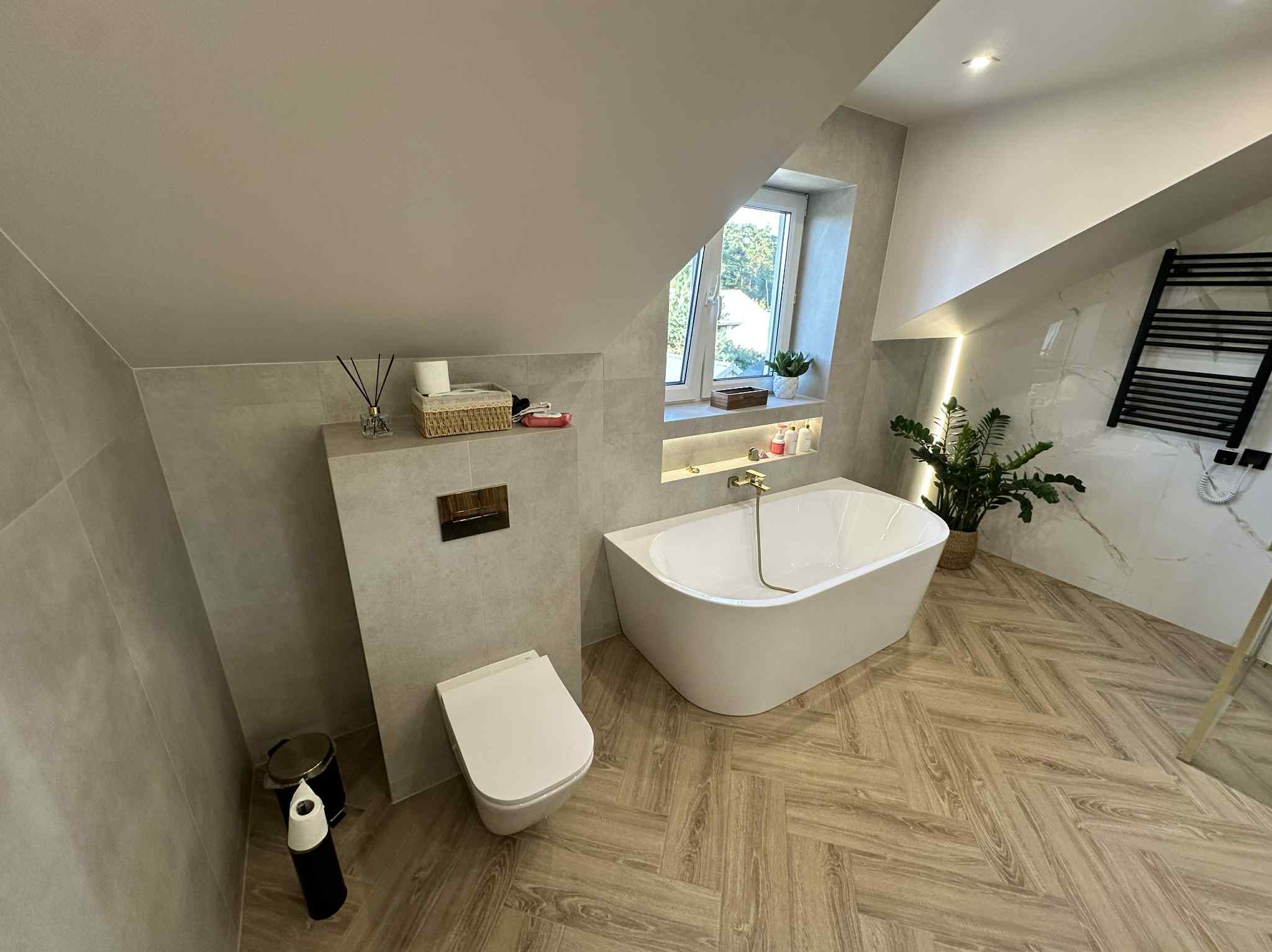 Nowoczesna łazienka na poddaszu z wolnostojącą wanną, toaletą podwieszaną i drewnopodobną podłogą ułożoną w jodełkę. Okno doświetla wnętrze.