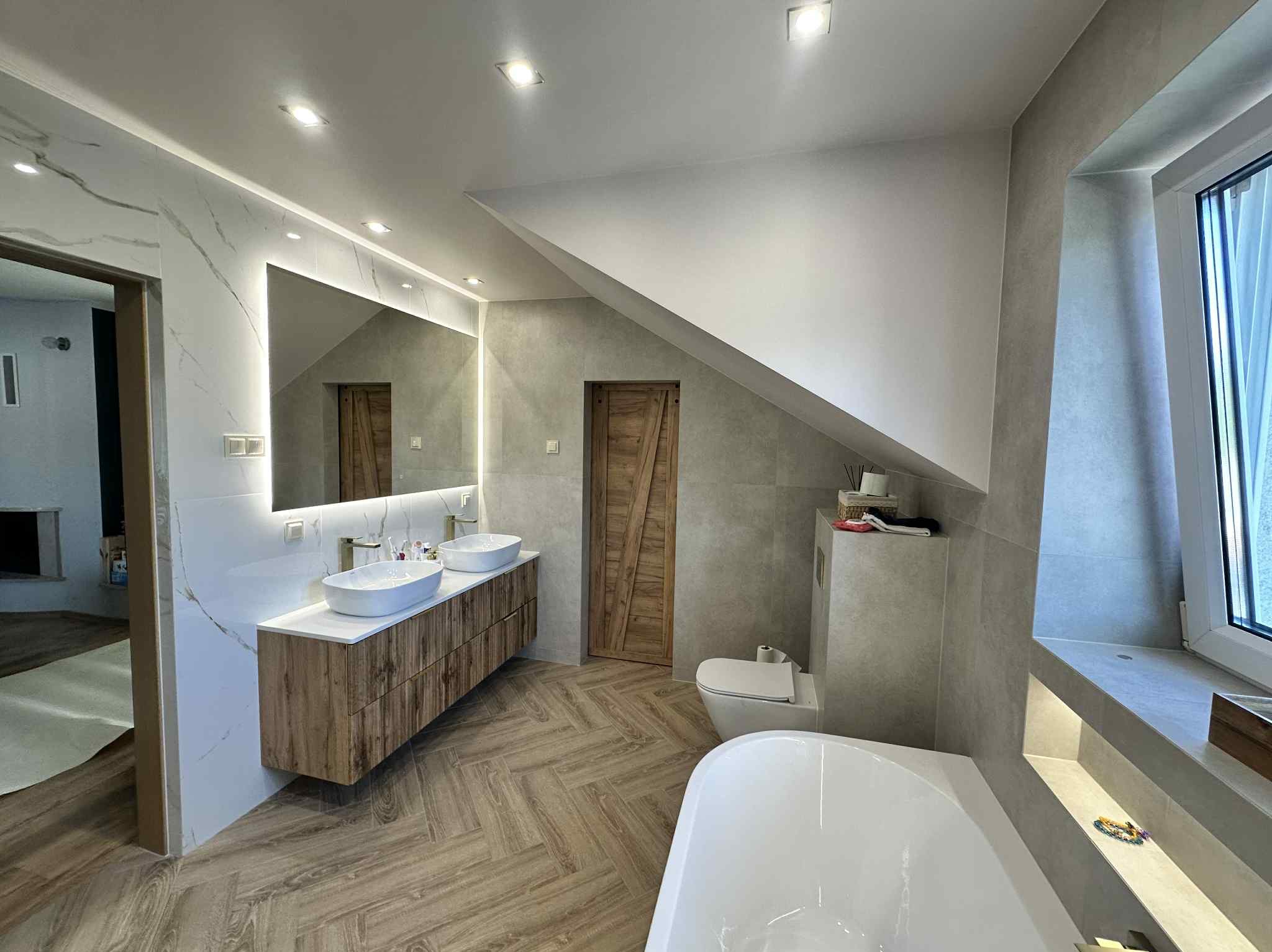 Nowoczesna łazienka na poddaszu z drewnianą podłogą w jodełkę, umywalkami nablatowymi i podświetlanym lustrem. Uwagę zwraca drewniana, rustykalna stolarka drzwi.