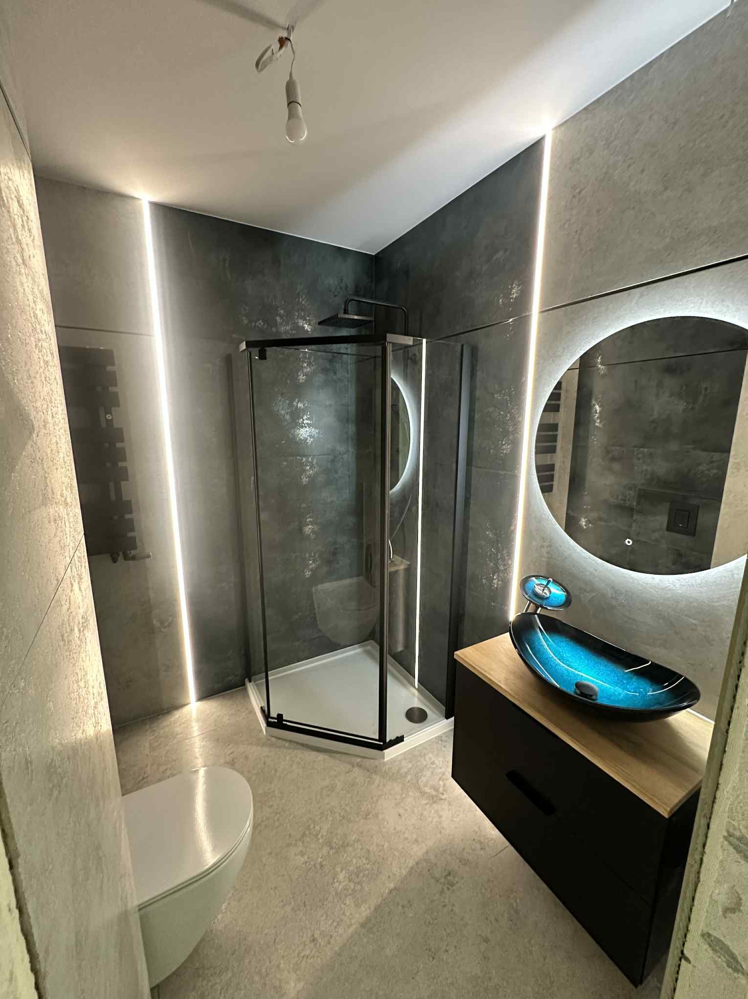 Nowoczesna łazienka w szarościach z podświetleniem LED, kabiną prysznicową typu walk-in i umywalką w kształcie łódki. Minimalistyczny design z okrągłym lustrem.