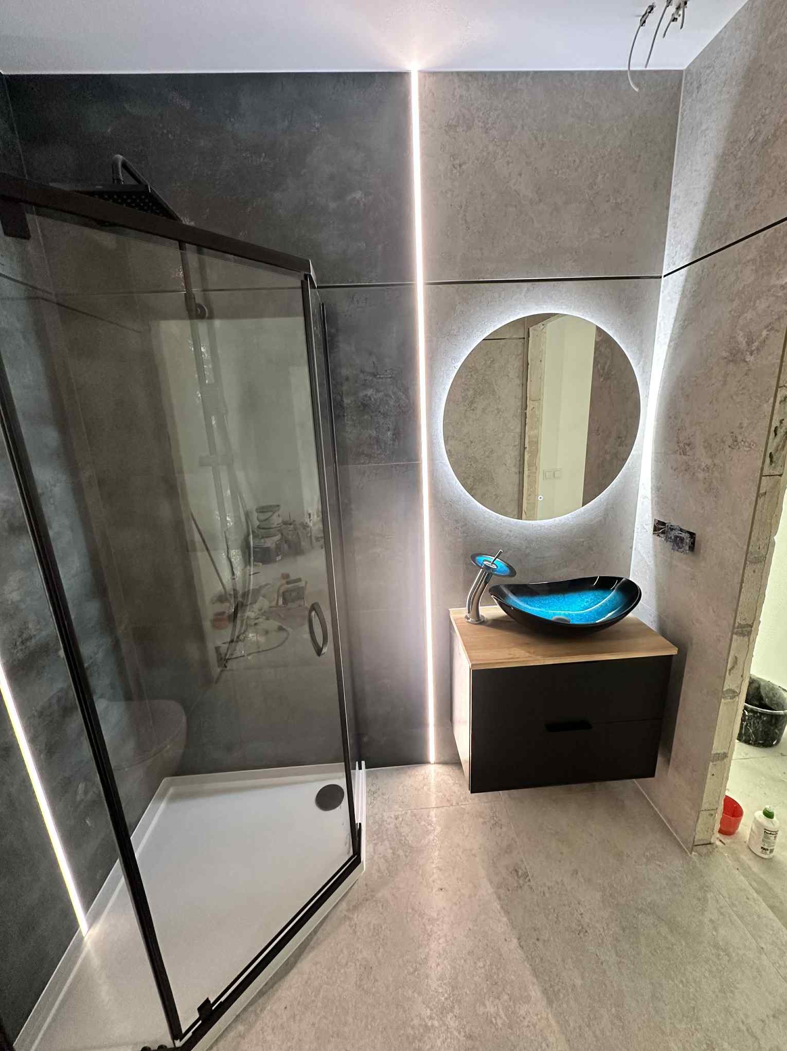 Nowoczesna łazienka z kabiną prysznicową typu walk-in, umywalką wpuszczaną w szafkę i okrągłym lustrem z podświetleniem LED. Szare płytki i oświetlenie liniowe.