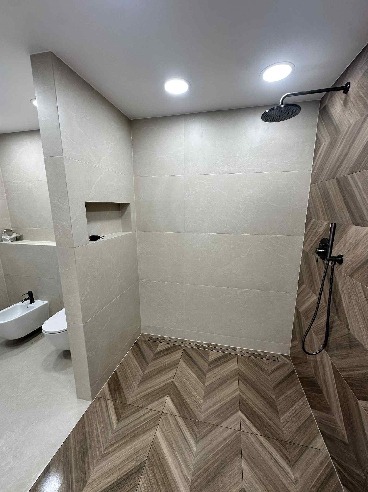 Nowoczesna łazienka z kabiną prysznicową typu walk-in, wykończona beżowymi i drewnopodobnymi płytkami ułożonymi w jodełkę. Minimalistyczna armatura w kolorze czarnym.