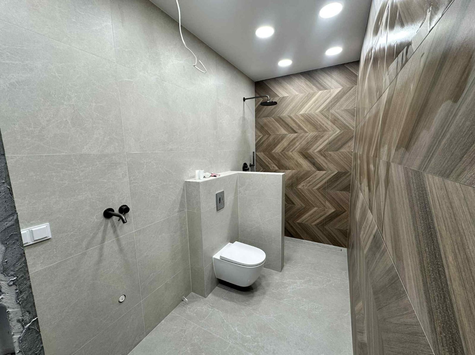 Nowoczesna łazienka z szarymi płytkami imitującymi marmur i akcentem z płytek drewnopodobnych ułożonych w jodełkę pod prysznicem. Czarna armatura i podwieszana toaleta.