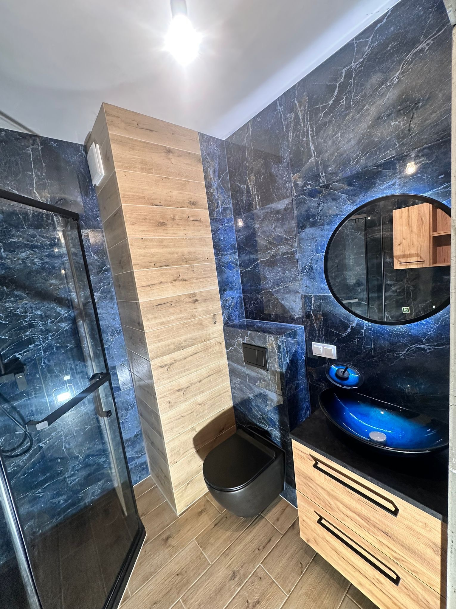 Stylowa łazienka z kabiną prysznicową, umywalką w odcieniach niebieskiego i drewnianymi akcentami. Marmurowe płytki w kolorze granatowym i beżowa podłoga.