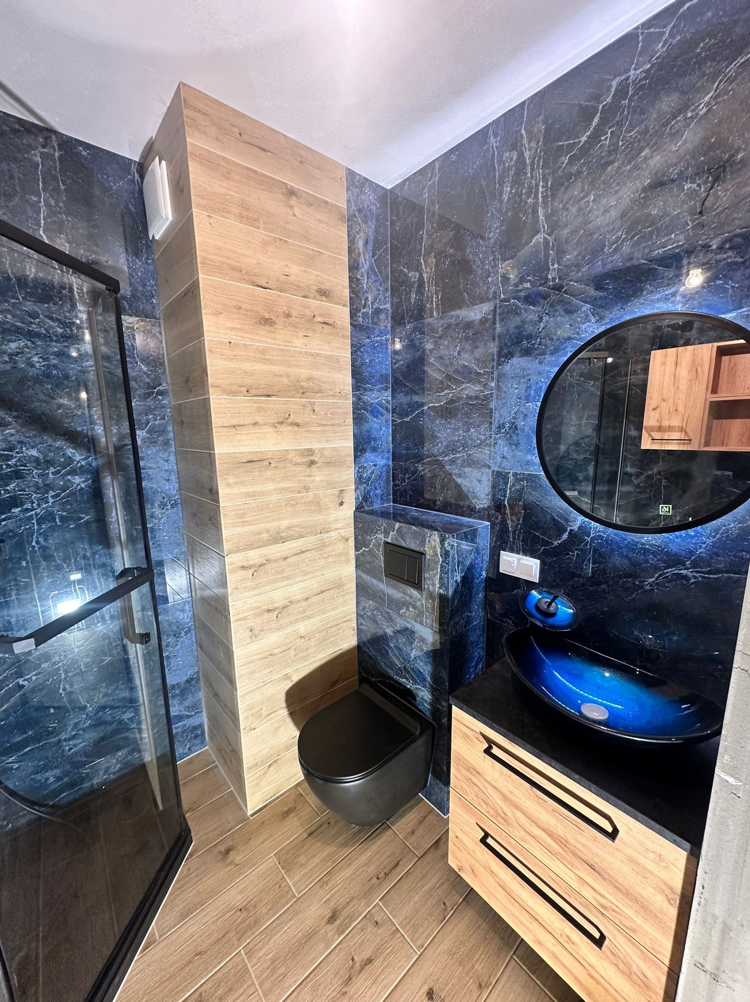 Nowoczesna łazienka z kabiną prysznicową, umywalką w odcieniach niebieskiego i toaletą w kolorze czarnym. Ściany wyłożone płytkami imitującymi marmur.